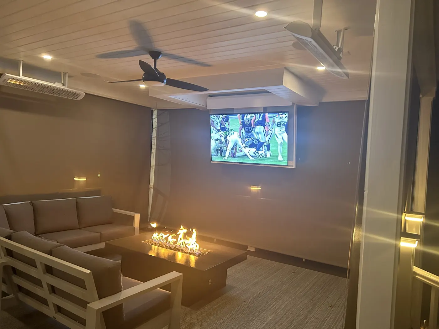 Enclosed outdoor room at night with overhead heaters, a ceiling fan, mounted television and a gas fire table