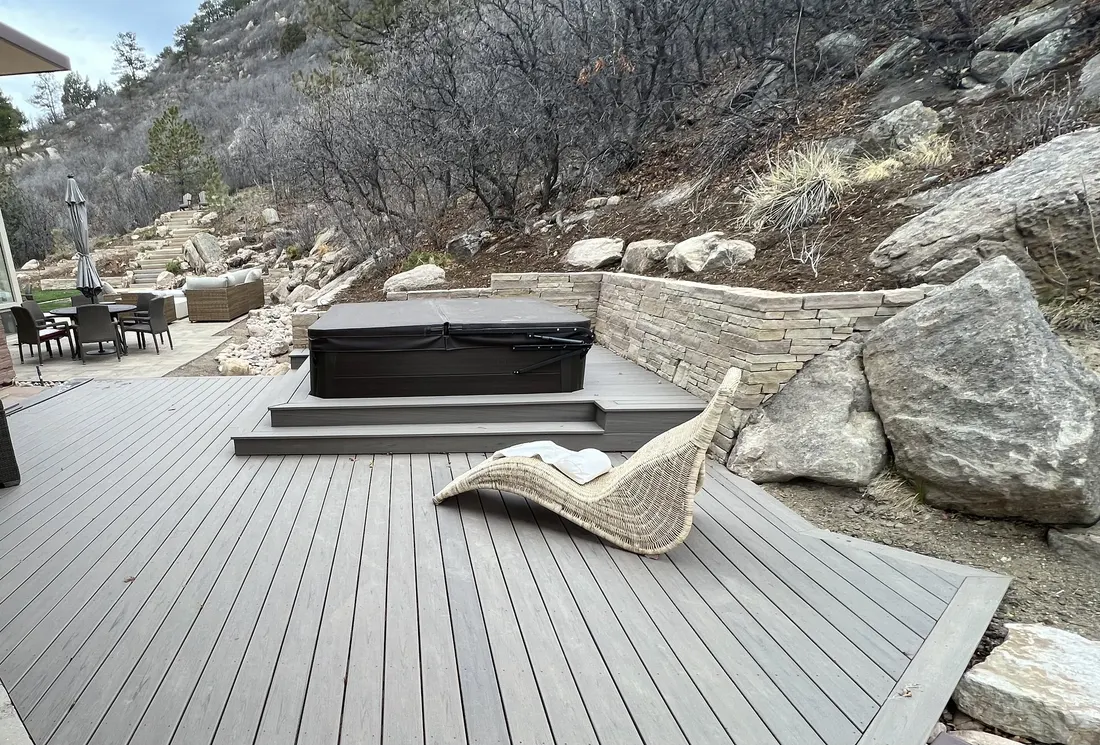 Composite deck stepping up to a raised spa, set against a stacked-stone retaining wall and boulders on a wooded slope