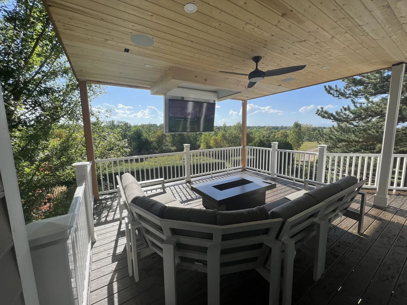 Covered deck with a tongue-and-groove ceiling, mounted television, fire table and white railing above a wooded view